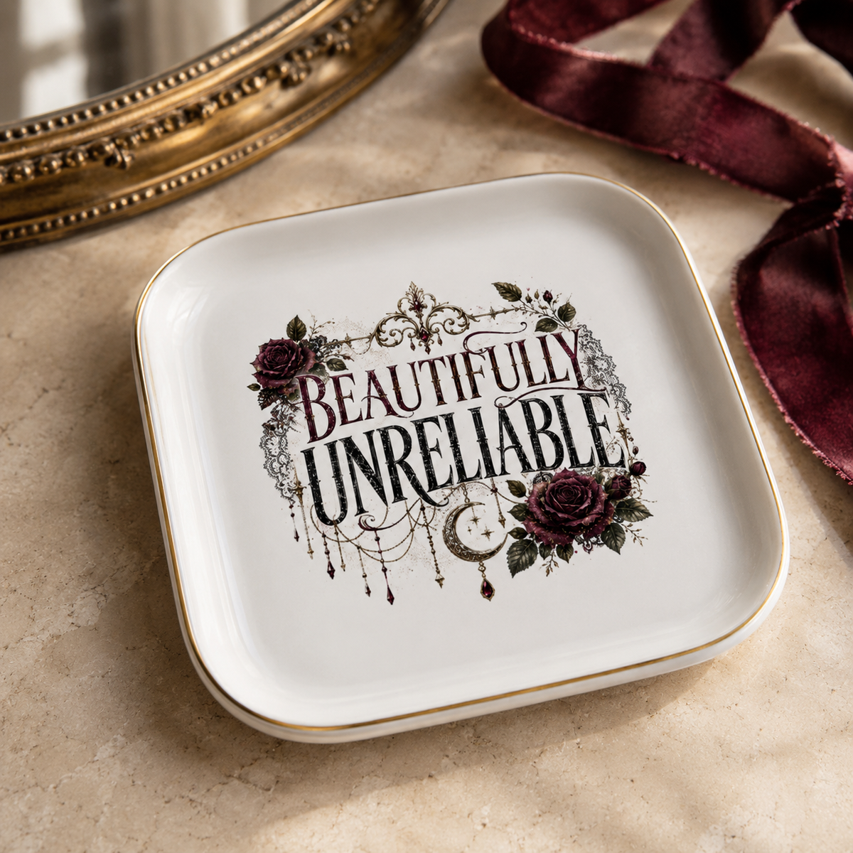 White gold-rimmed “Beautifully Unreliable” jewelry dish with burgundy roses, crescent moon, ornate filigree and dangling jewel motifs, beside a gold mirror and burgundy ribbon.