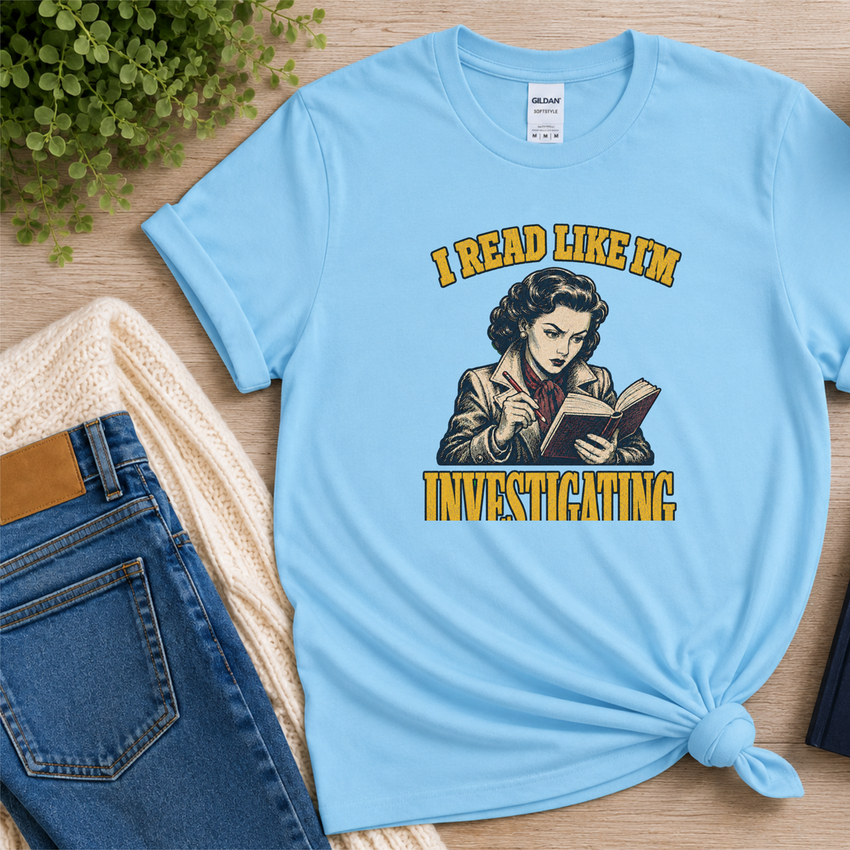 Light blue T-shirt laid flat with an illustrated reader holding a book and pen and the words “I Read Like I’m Investigating.”