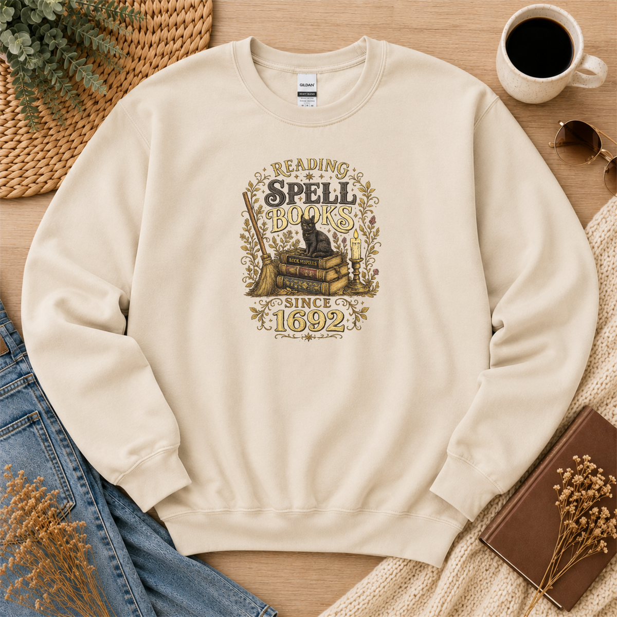 Sand crewneck sweatshirt laid flat with a black cat on stacked books with candles and botanical flourishes and the words “Reading Spell Books Since 1692.”