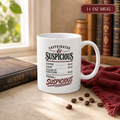 11 oz white “Caffeinated & Suspicious” receipt-style mug on a tabletop beside a mystery novel, burgundy cloth, and coffee beans.