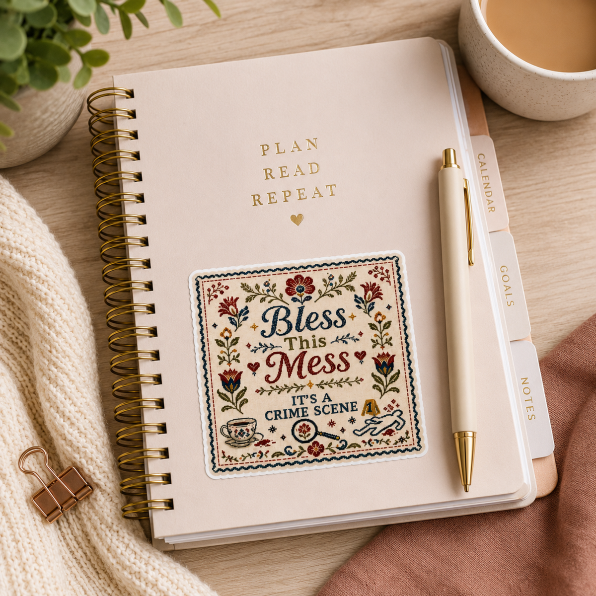 “Bless This Mess. It’s a Crime Scene” sticker with a folk-art floral border, teacup, magnifying glass, evidence marker, and body outline, shown on a cream spiral planner.