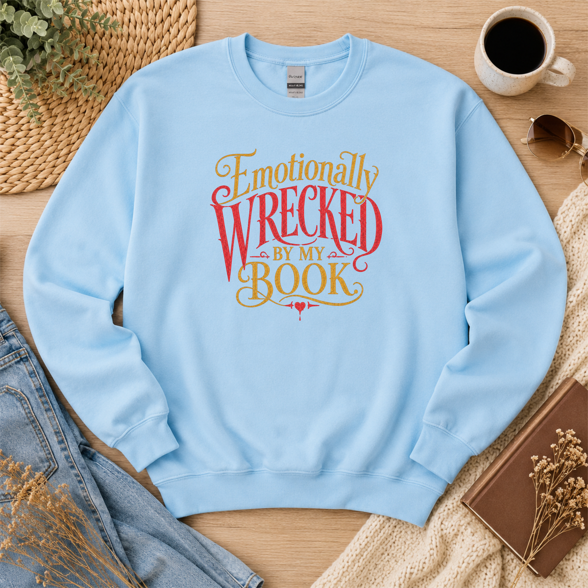 Light blue crewneck sweatshirt laid flat with ornate red and gold lettering and the words “Emotionally Wrecked by My Book.”