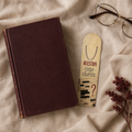 Cream aluminum page-holder bookmark reading “Question Every Chapter,” with redacted notes, circled clues, and a red question mark, on linen beside a burgundy book and glasses.