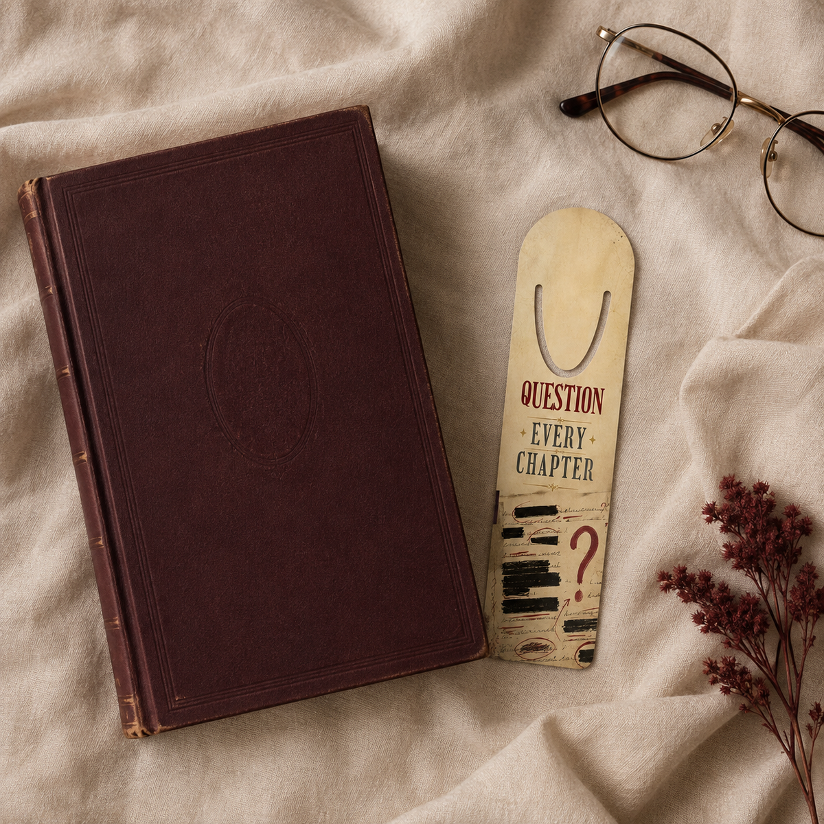 Cream aluminum page-holder bookmark reading “Question Every Chapter,” with redacted notes, circled clues, and a red question mark, on linen beside a burgundy book and glasses.