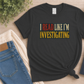 Black T-shirt laid flat with bold vintage lettering and the words “I Read Like I’m Investigating.”