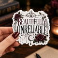 Hand holding a white-bordered sticker reading “Beautifully Unreliable,” featuring burgundy and black gothic lettering, dark roses, lace-like flourishes, dangling ornaments, and a crescent moon.