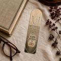 Cream aluminum page-holder bookmark with oversized fingerprint-pattern lines and the words “Reading Between the Lies,” on linen beside an old book, glasses, and dried flowers.