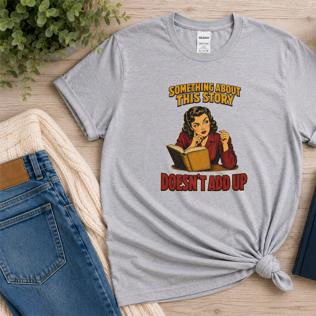 Sport gray T-shirt laid flat with an illustrated reader holding an open book and the words “Something About This Story Doesn’t Add Up.”