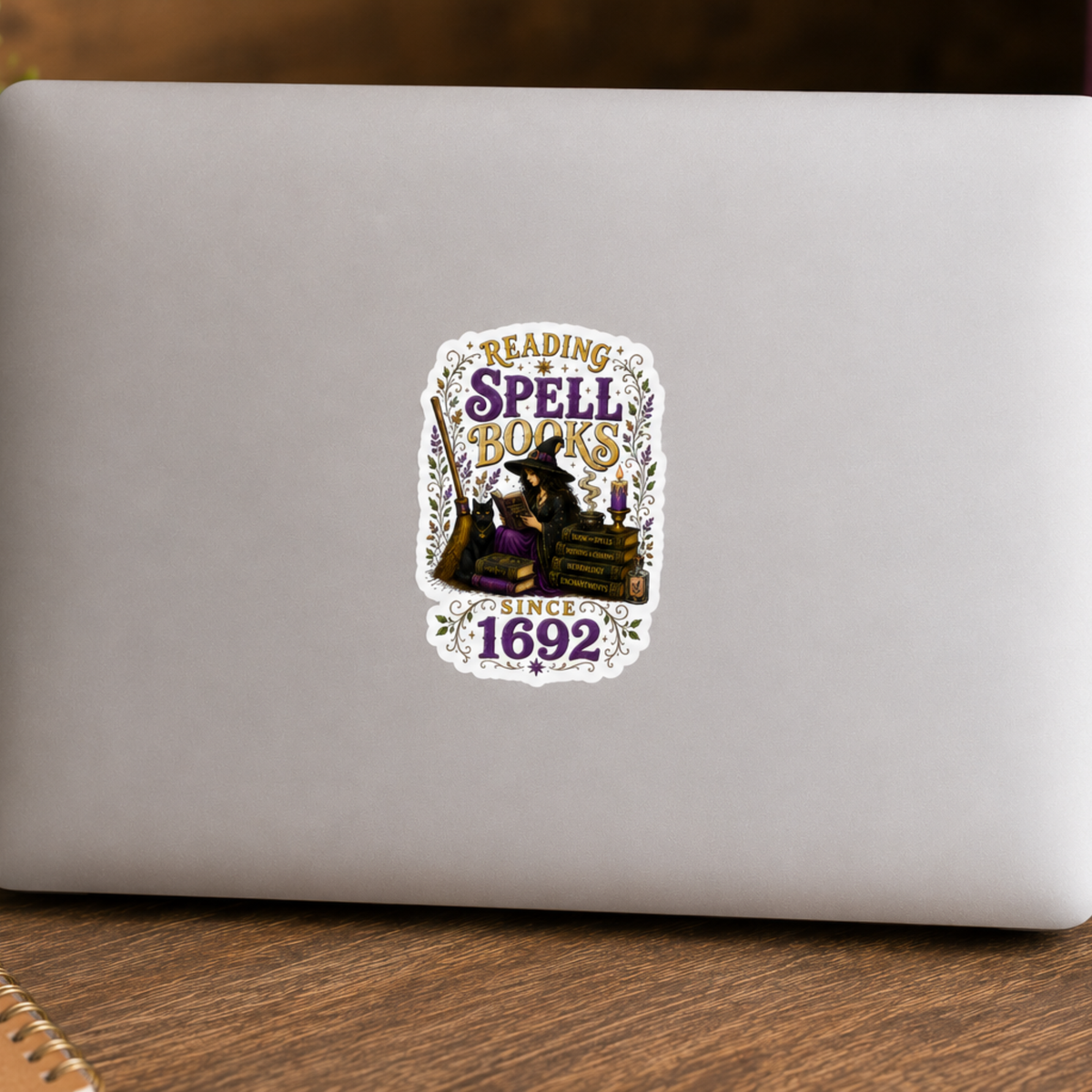 “Reading Spell Books Since 1692” sticker with purple and gold lettering, a witch reading beside a black cat, candle, broom, and stacked spell books, shown on a silver laptop.