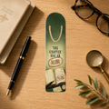 Green aluminum page-holder bookmark reading “This Chapter Has an Alibi,” with open books stamped “Alibi,” on a wooden desk beside a reading journal, fountain pen, glasses, and magnifying glass.