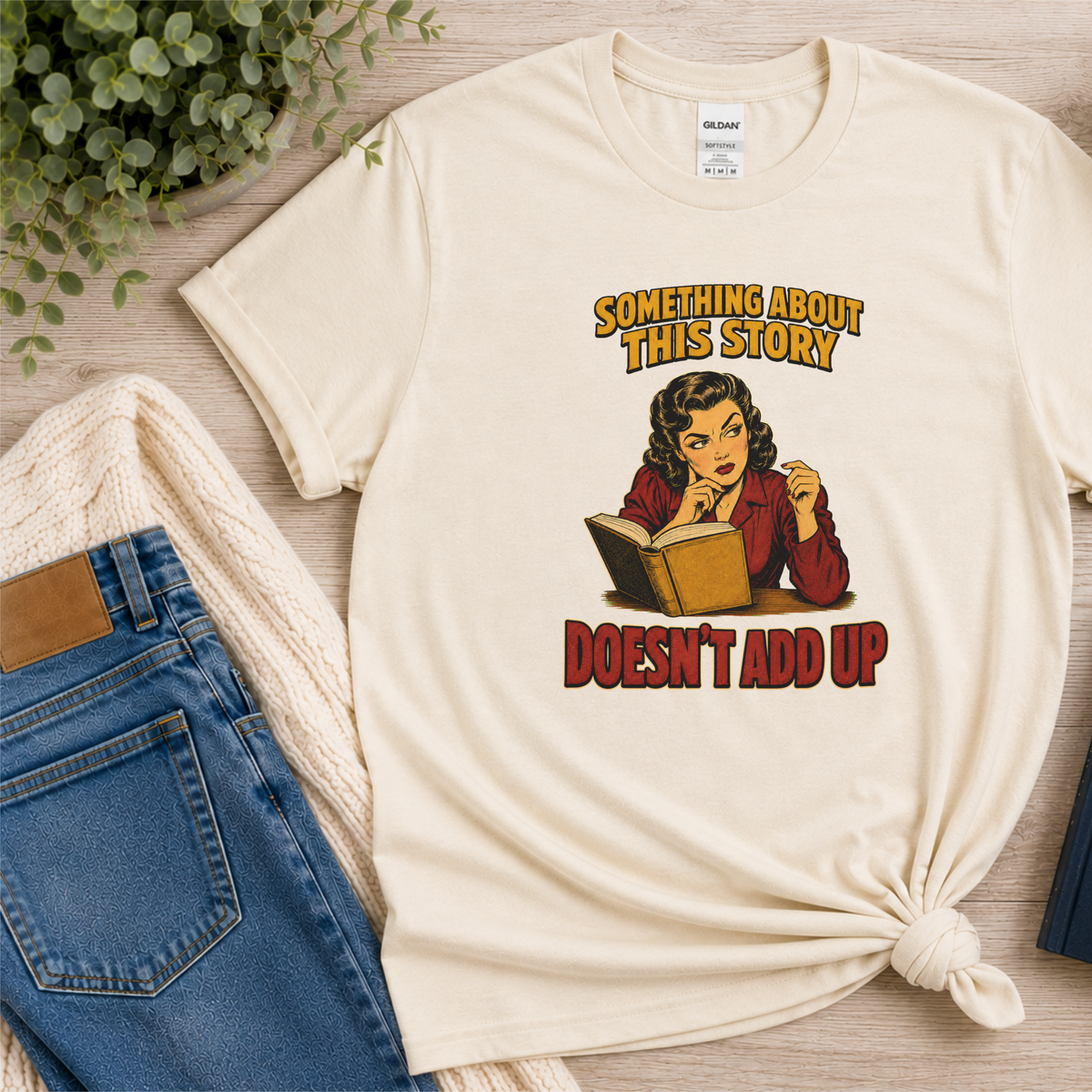 Natural cream T-shirt laid flat with an illustrated reader holding an open book and the words “Something About This Story Doesn’t Add Up.”