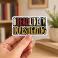 Hand holding a white-bordered sticker reading “I Read Like I’m Investigating,” featuring red, cream, and gold block lettering.