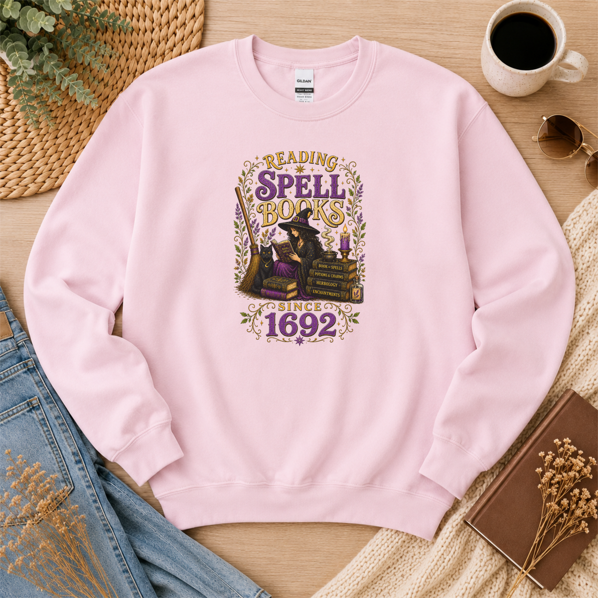 Light pink crewneck sweatshirt laid flat with a witch reading beside stacked books and candles and the words “Reading Spell Books Since 1692.”