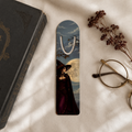 Aluminum page-holder bookmark featuring a witch in a dark red gown and pointed hat beneath a pale full moon and bare branches, on cream fabric beside a gothic book and glasses.