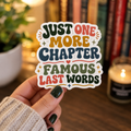 Hand holding a white-bordered sticker reading “Just One More Chapter. Famous Last Words,” featuring green, coral, gold, and blue retro lettering with hearts and sparkles.