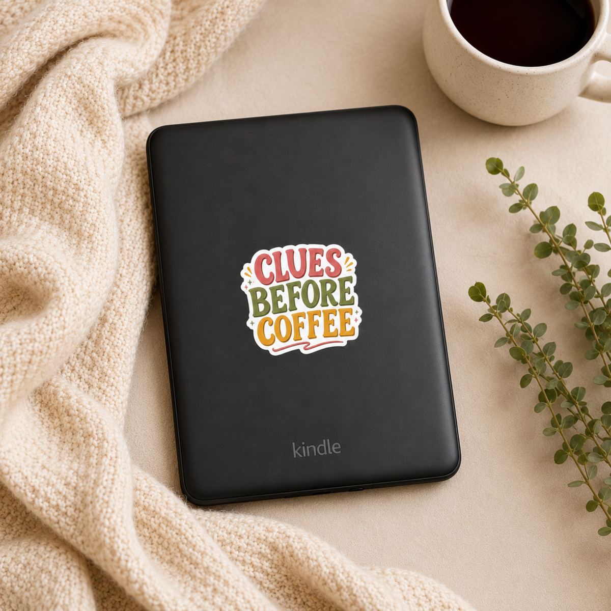 “Clues Before Coffee” sticker with coral, olive, and gold block lettering with small sparkles, shown on a black Kindle.
