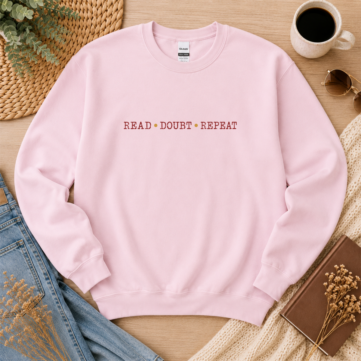 Light pink crewneck sweatshirt laid flat with small red lettering across the chest and the words “Read Doubt Repeat.”