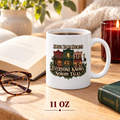 11-ounce Small Town Secrets mug featuring illustrated storefronts and the phrase “Everyone Knows. Nobody Talks.” beside an open book, eyeglasses, candle and mystery novel.