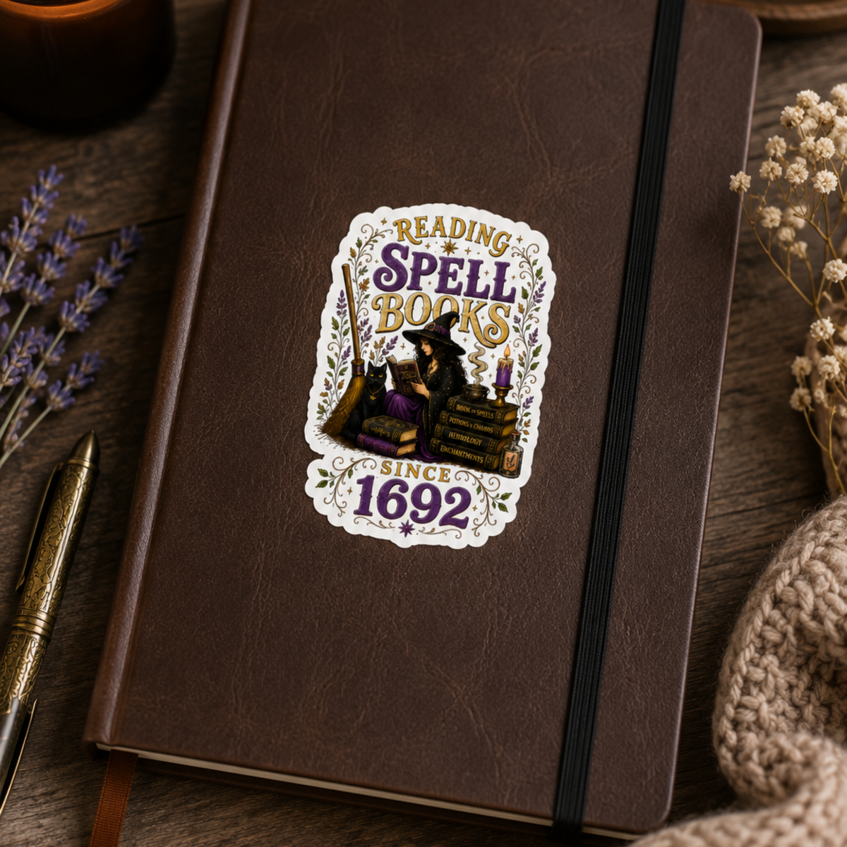 “Reading Spell Books Since 1692” sticker with purple and gold lettering, a witch reading beside a black cat, candle, broom, and stacked spell books, shown on a brown notebook.