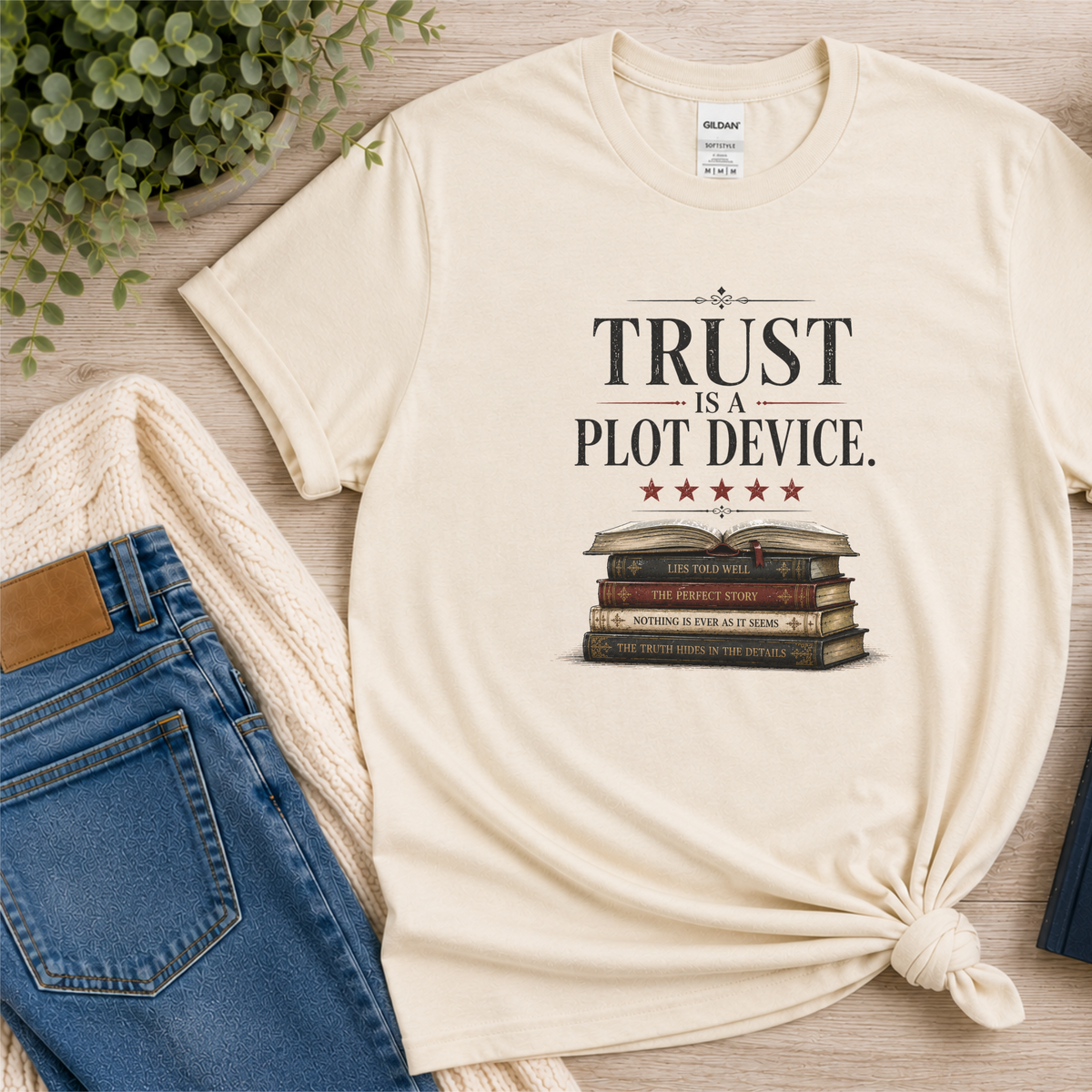 Natural cream T-shirt laid flat with stars above a stack of books and the words “Trust Is a Plot Device.”