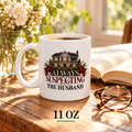 White 11 oz mug of coffee beside an open book, glasses and flowers, with a house, red roses and “Always Suspecting the Husband” in gold, burgundy and black lettering.