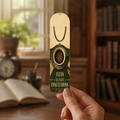 Hand holding a cream-and-dark-green aluminum page-holder bookmark featuring a fingerprint in a coffee mug and the words “Clues Before Conclusions” in a library.