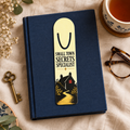 Gold-toned aluminum page-holder bookmark reading “Small Town Secrets Specialist,” with a winding path to a black house with a red-lit window, on a navy book.