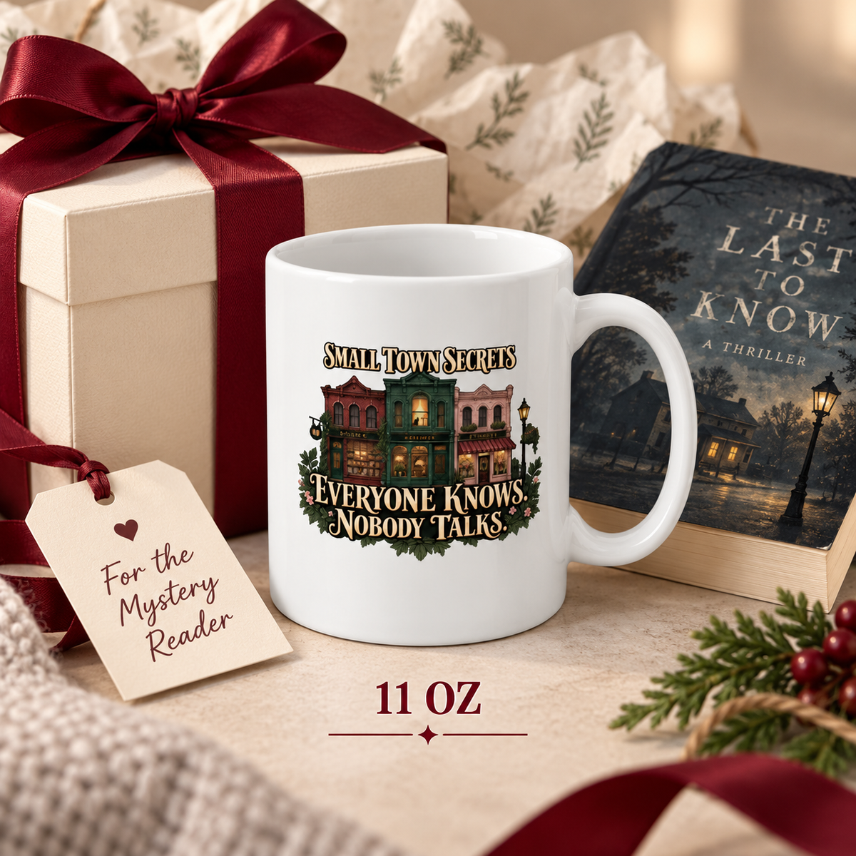 11-ounce Small Town Secrets mug beside a cream gift box with burgundy bow, mystery-reader gift tag, evergreen branches and a thriller titled “The Last to Know.”