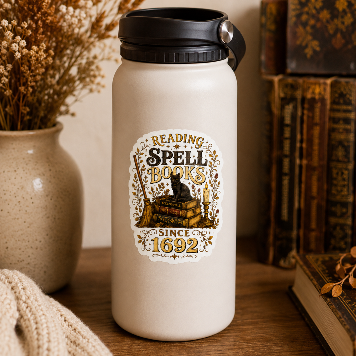 “Reading Spell Books Since 1692” sticker with black and gold lettering, a black cat on antique books, a broom, candle, and botanical flourishes, shown on a cream water bottle.