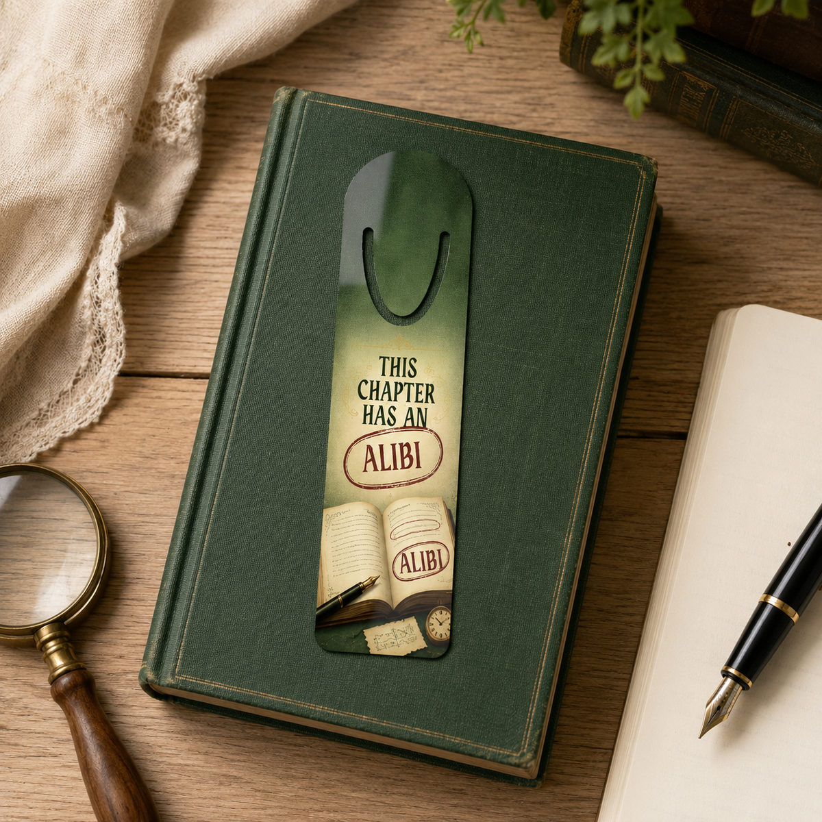 “This Chapter Has an Alibi” bookmark on a green clothbound book beside a magnifying glass, fountain pen, and open notebook.