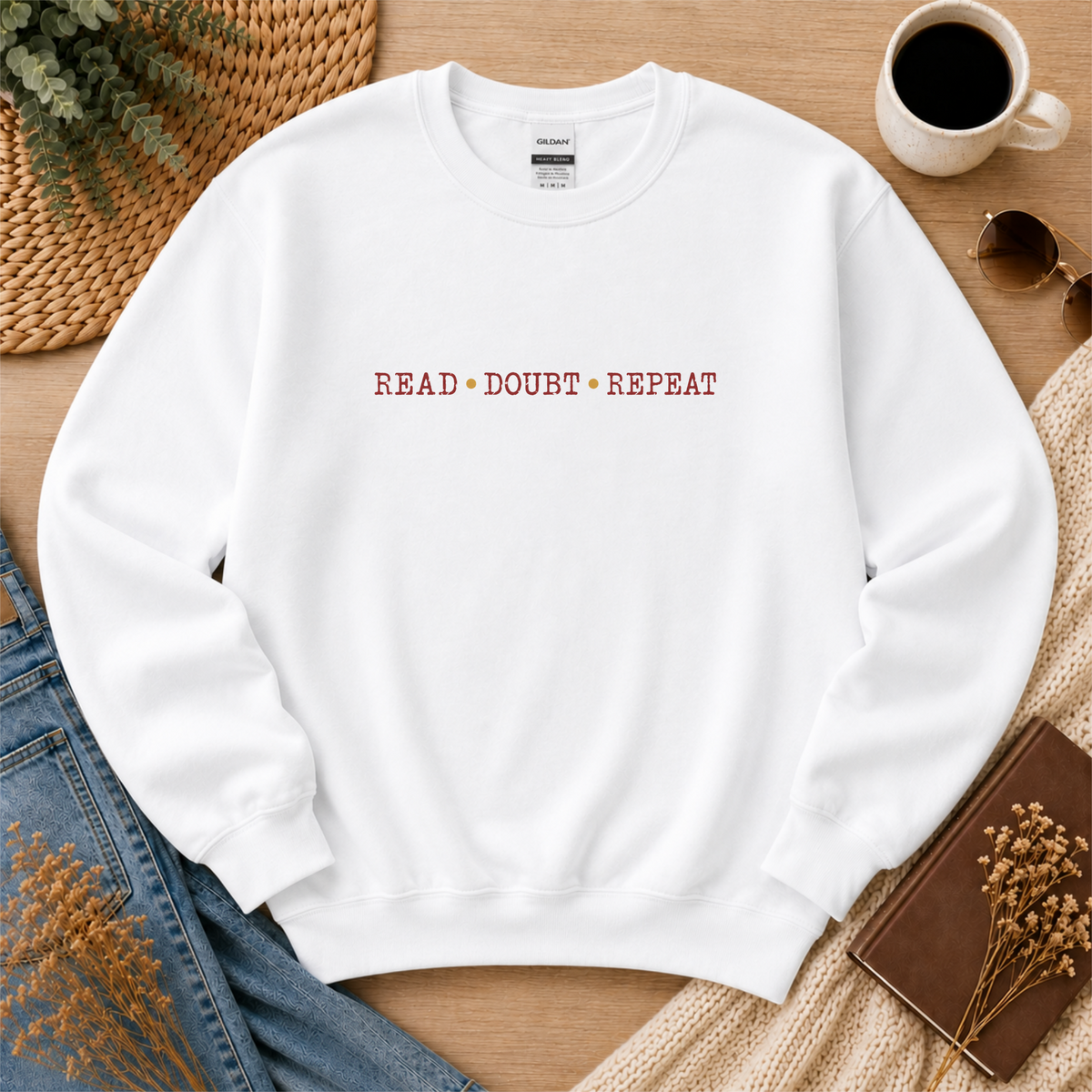 White crewneck sweatshirt laid flat with small red lettering across the chest and the words “Read Doubt Repeat.”
