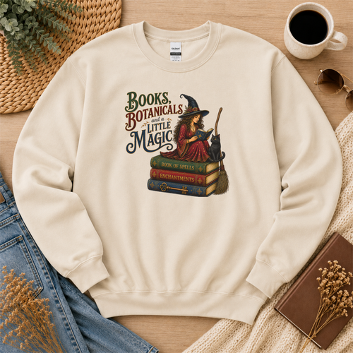 Sand crewneck sweatshirt laid flat with a witch reading on stacked spell books beside a black cat and broom and the words “Books, Botanicals and a Little Magic.”