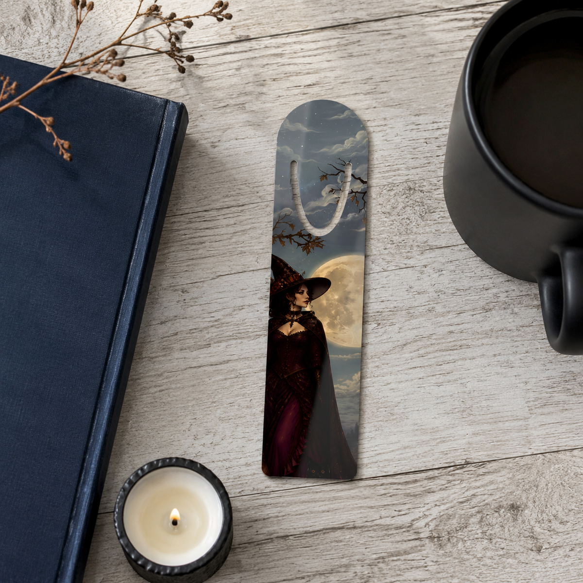 Aluminum bookmark showing a witch beneath a full moon on a pale wooden table beside a navy book, black mug, and lit candle.