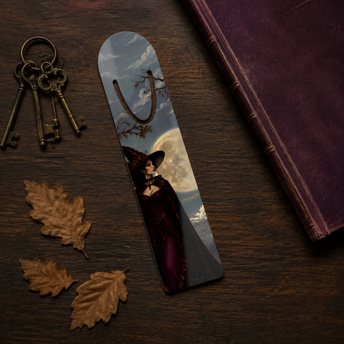 Witch-and-full-moon bookmark on dark wood beside antique-style keys, burgundy book, and dried autumn leaves.