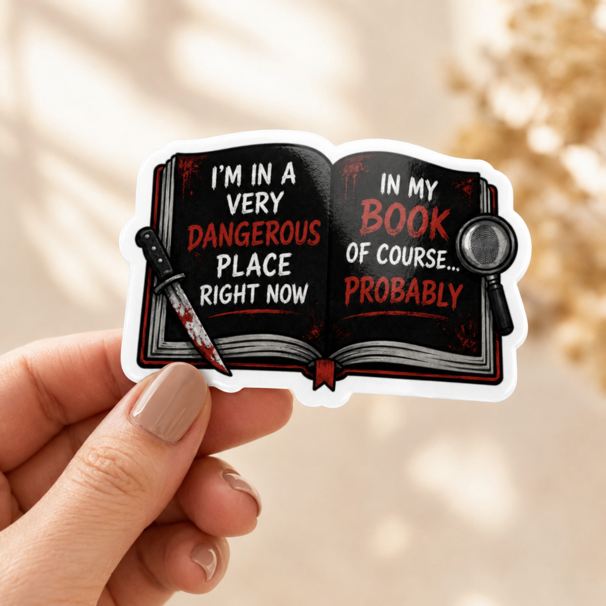 Hand holding a white-bordered sticker reading “I’m in a very dangerous place right now. In my book, of course… probably,” featuring an open black book with red and white lettering, a bloodstained knife, and a magnifying glass.