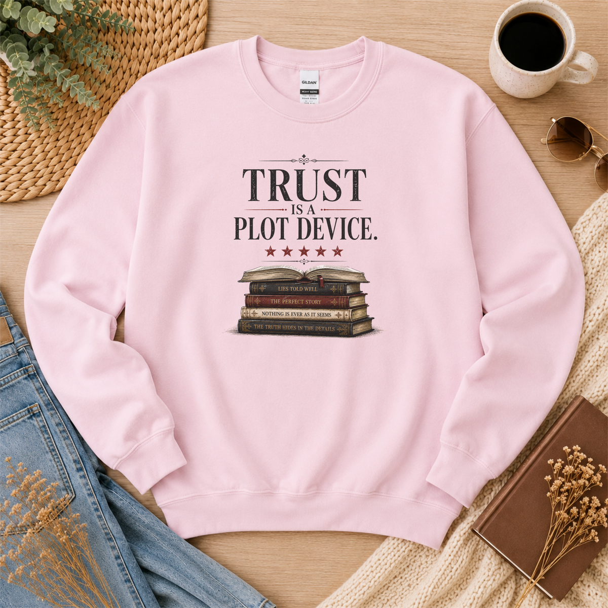 Light pink crewneck sweatshirt laid flat with stars above a stack of books and the words “Trust Is a Plot Device.”