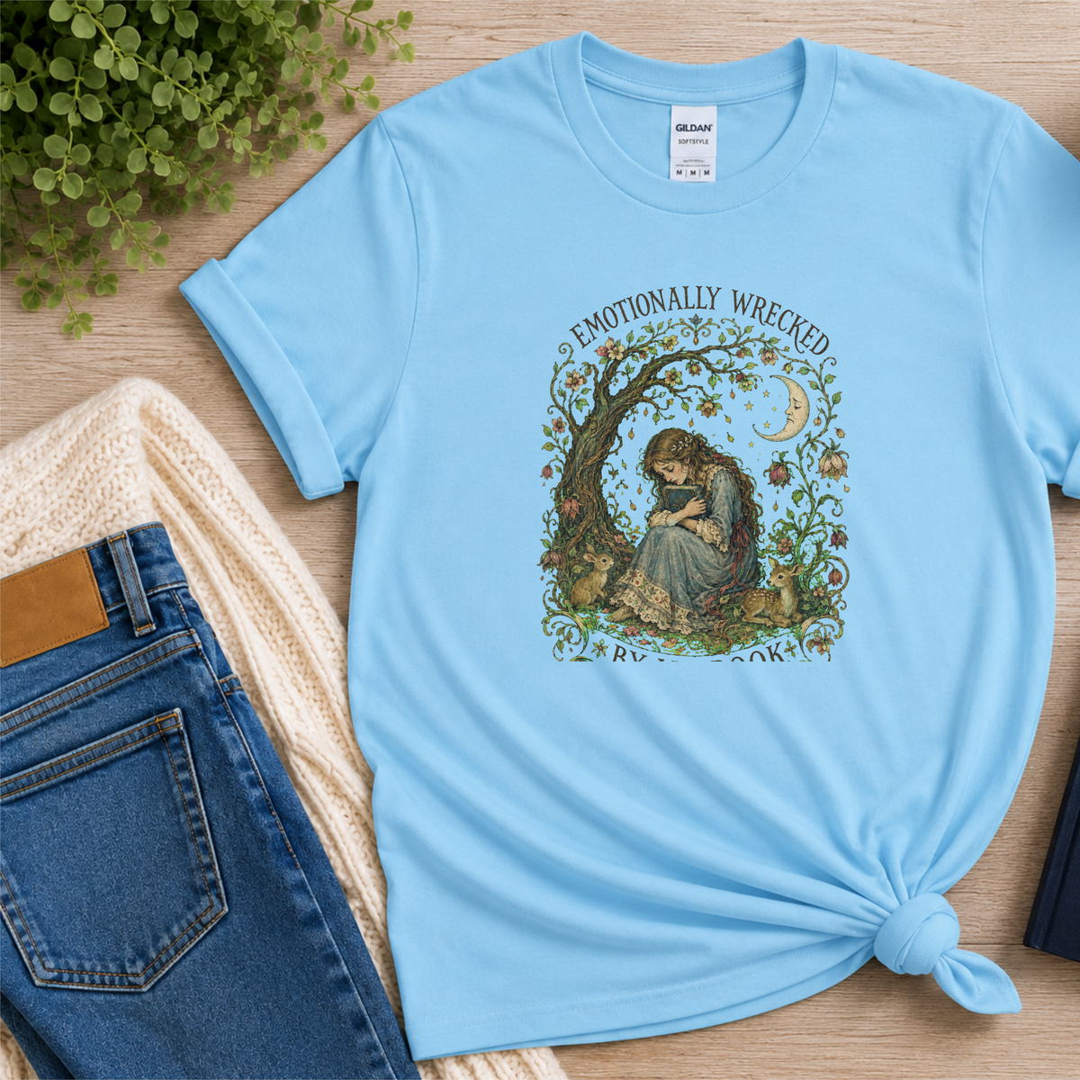 Light blue T-shirt laid flat with a reader beneath a tree, flowers and a crescent moon and the words “Emotionally Wrecked by My Book.”