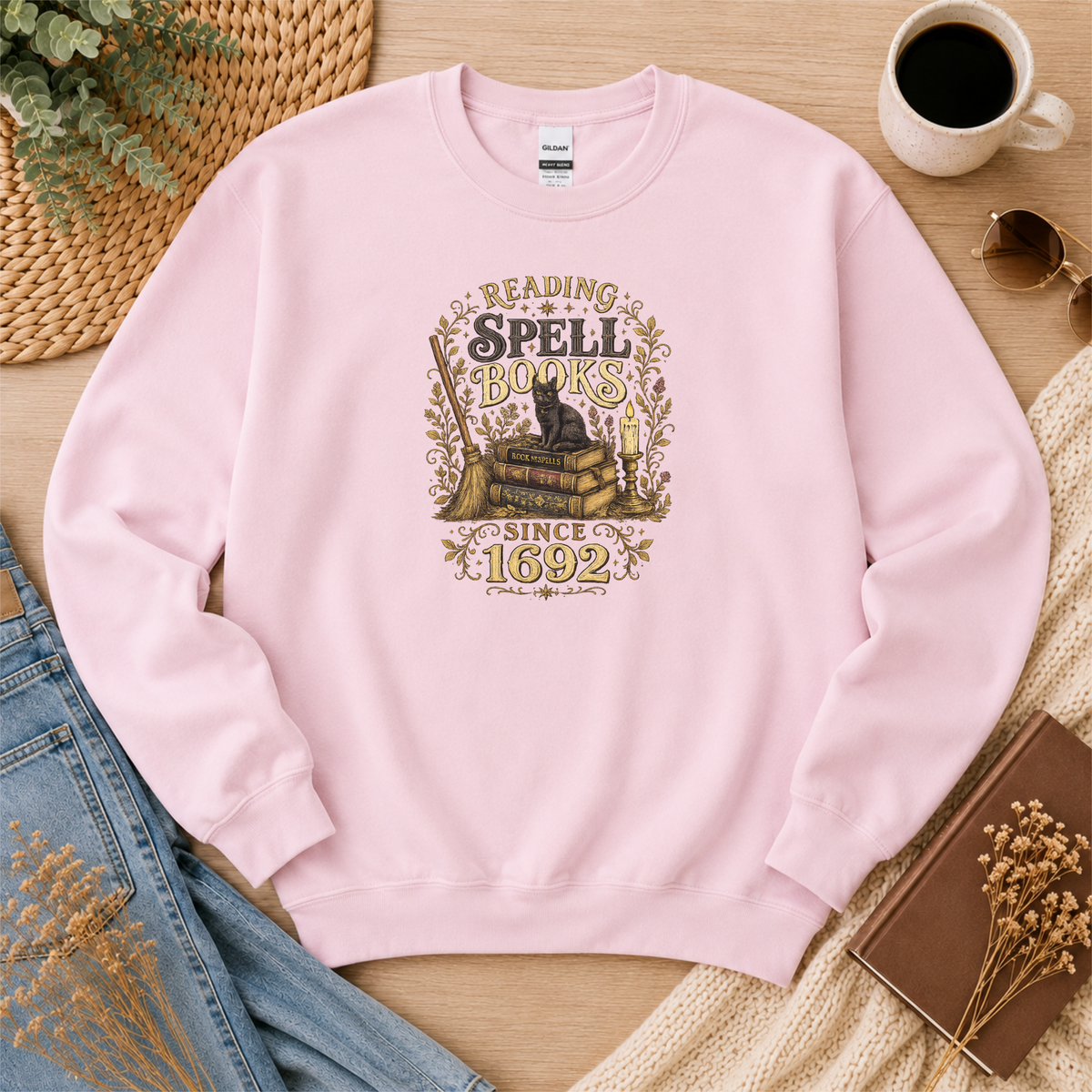 Light pink crewneck sweatshirt laid flat with a black cat on stacked books with candles and botanical flourishes and the words “Reading Spell Books Since 1692.”
