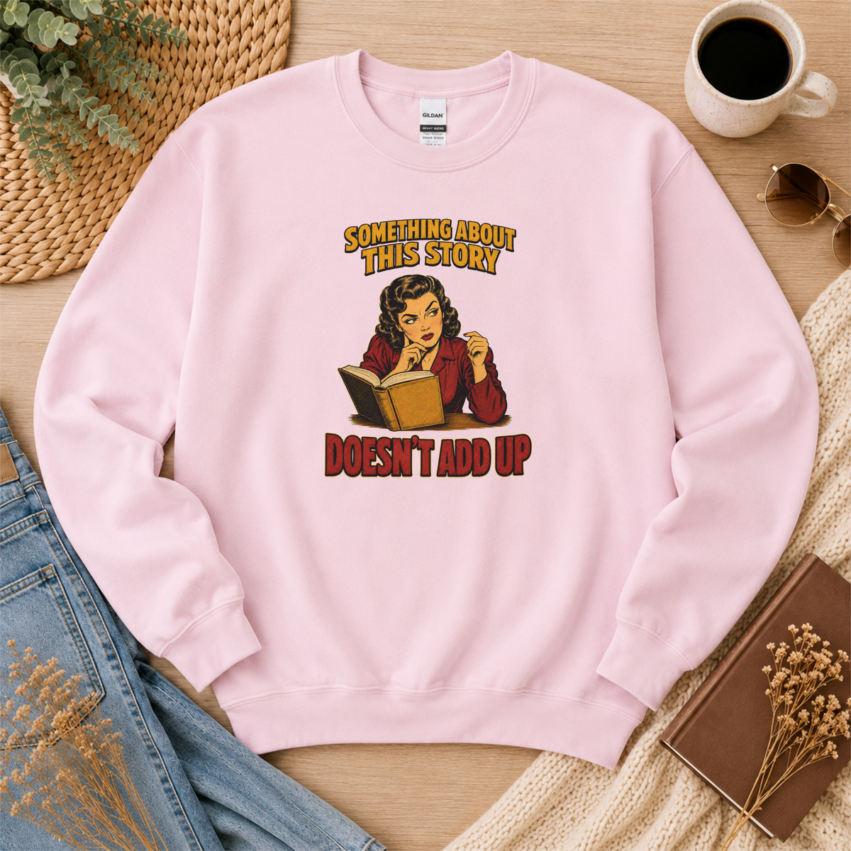 Light pink crewneck sweatshirt laid flat with an illustrated reader holding an open book and the words “Something About This Story Doesn’t Add Up.”