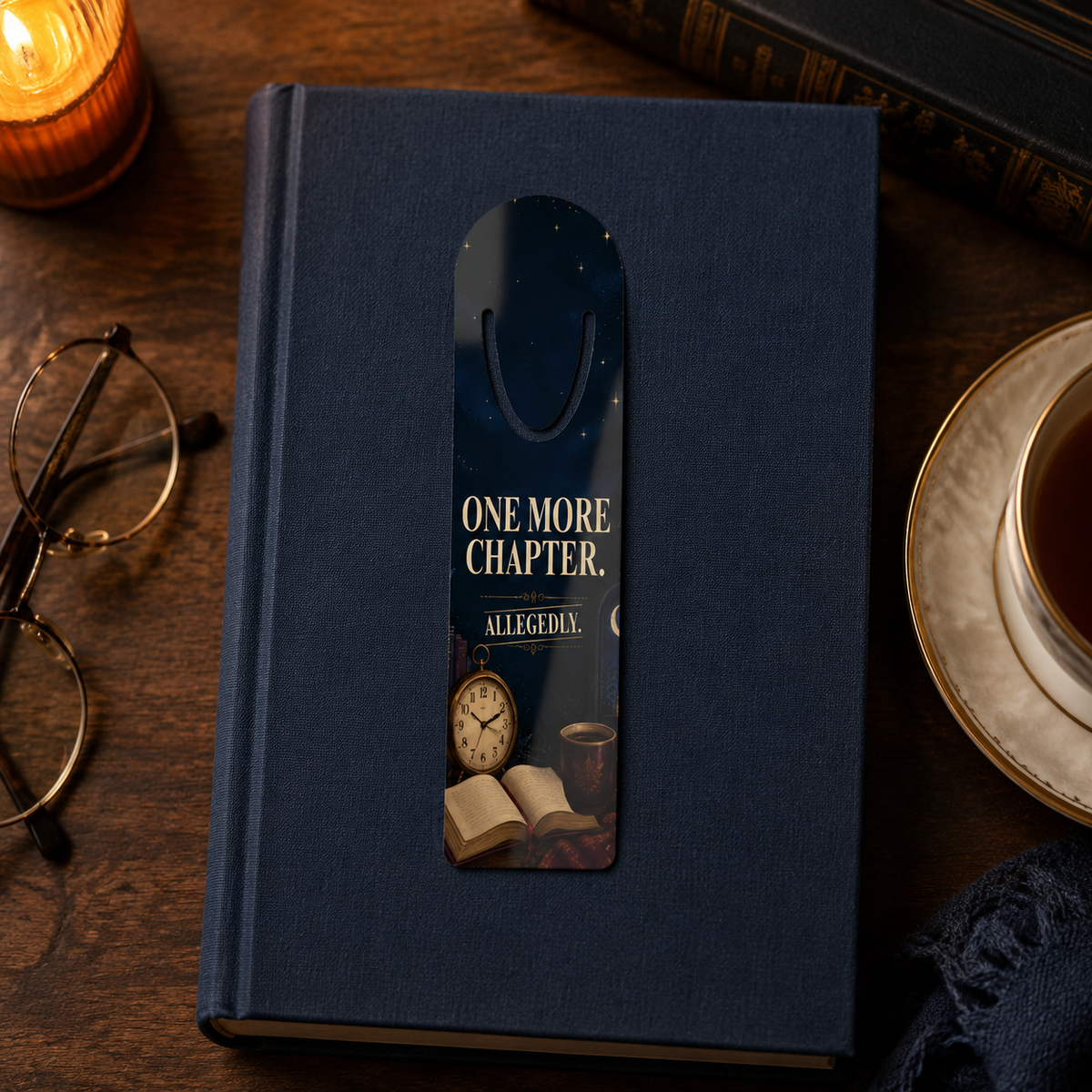 “One More Chapter. Allegedly.” bookmark resting on a navy clothbound book beside reading glasses, tea, and a glowing candle.