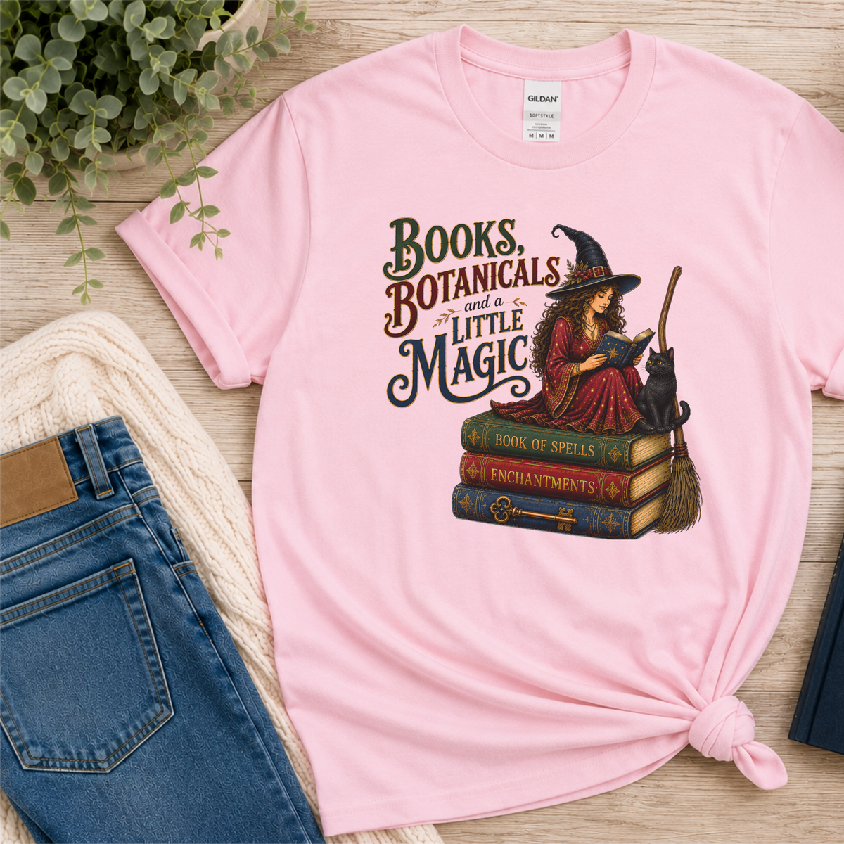 Light pink T-shirt laid flat with a witch reading on stacked spell books beside a black cat and broom and the words “Books, Botanicals and a Little Magic.”