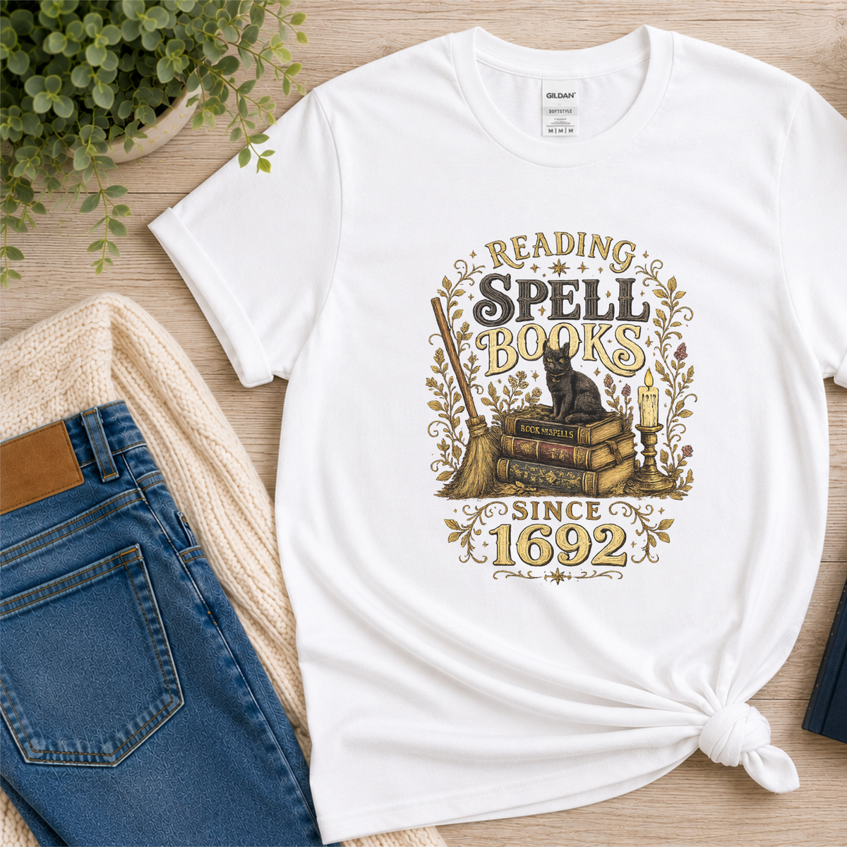 White T-shirt laid flat with a black cat on stacked books with candles and botanical flourishes and the words “Reading Spell Books Since 1692.”