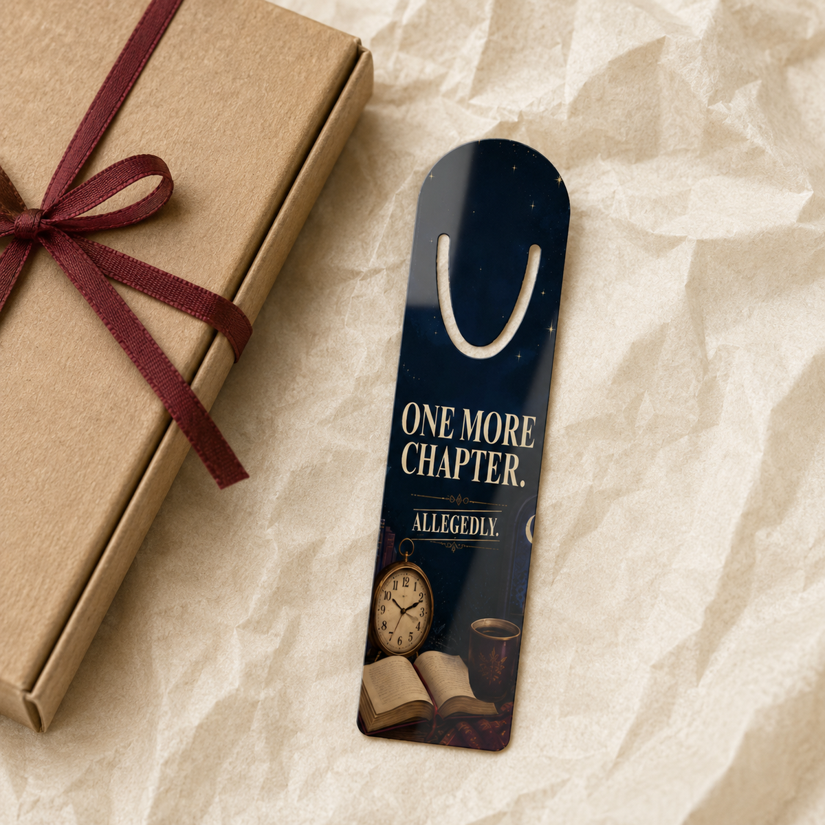 Navy aluminum page-holder bookmark reading “One More Chapter. Allegedly.” above an antique clock, open book, and warm drink, beside a kraft gift tied with burgundy ribbon.