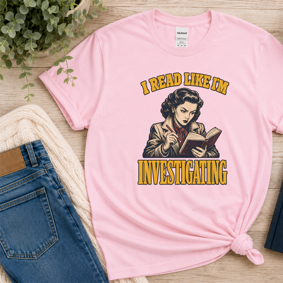 Light pink T-shirt laid flat with an illustrated reader holding a book and pen and the words “I Read Like I’m Investigating.”