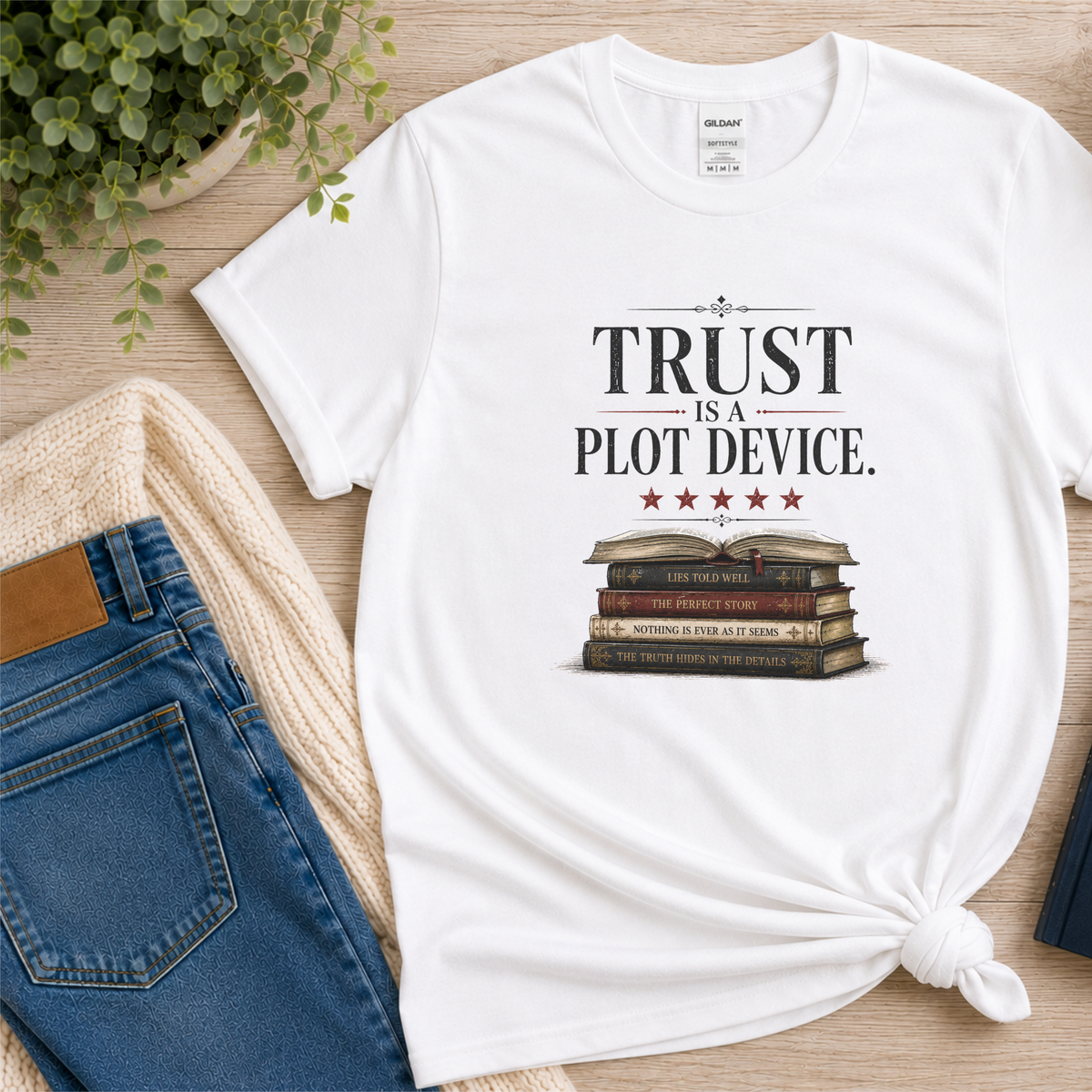 White T-shirt laid flat with stars above a stack of books and the words “Trust Is a Plot Device.”