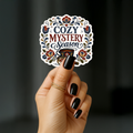 Hand holding a white-bordered sticker reading “Cozy Mystery Season,” featuring navy and burgundy lettering, a floral folk-art border, decorated teacup, and magnifying glass.