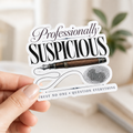Hand holding a white-bordered sticker reading “Professionally Suspicious. Trust No One • Question Everything,” featuring black lettering, a fountain pen, ink flourishes, and a fingerprint.