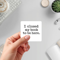 Hand holding a white-bordered sticker reading “I closed my book to be here,” featuring black typewriter lettering on a white rounded rectangle.