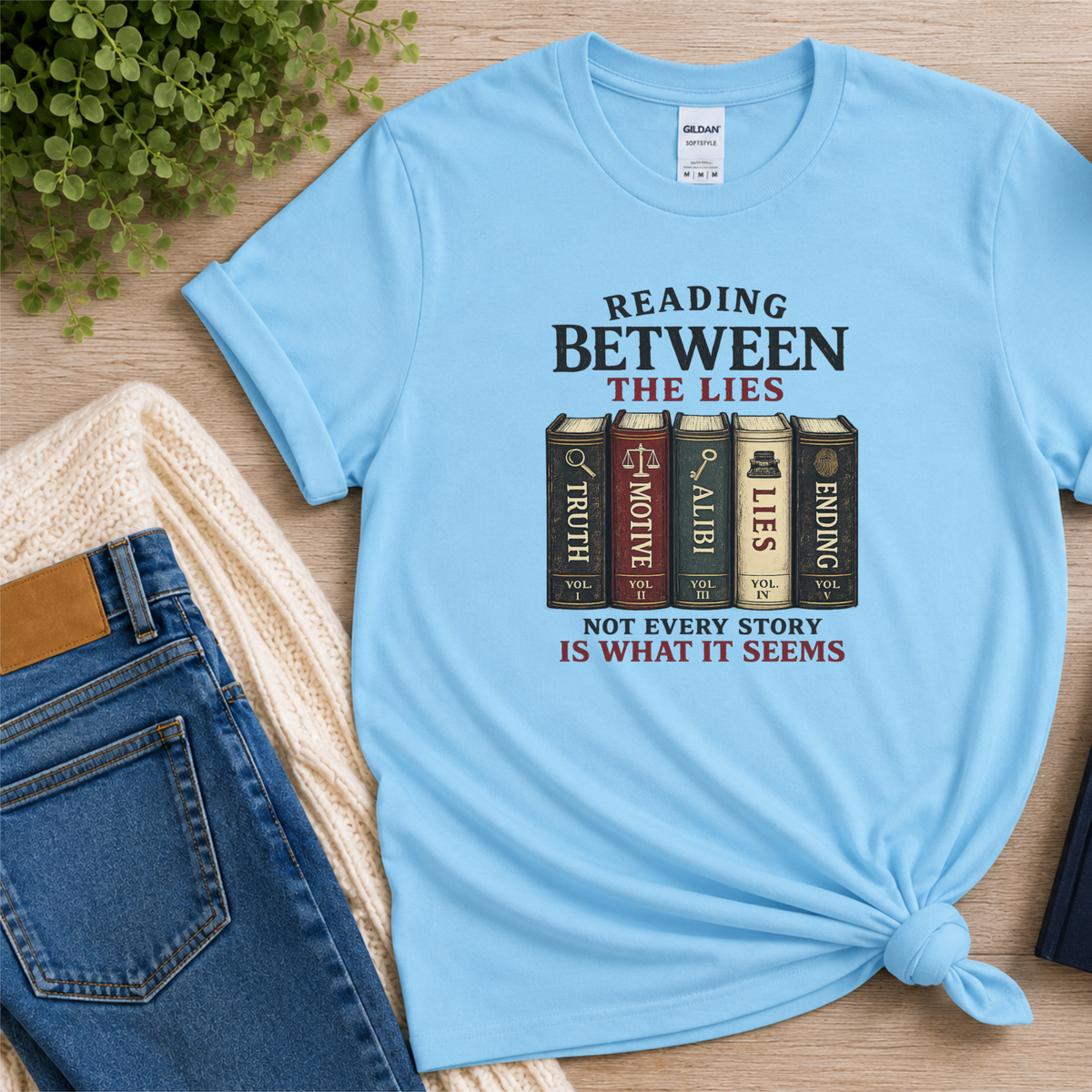 Light blue T-shirt laid flat with books labeled Truth, Motive, Alibi, Lies and Ending and the words “Reading Between the Lies. Not Every Story Is What It Seems.”