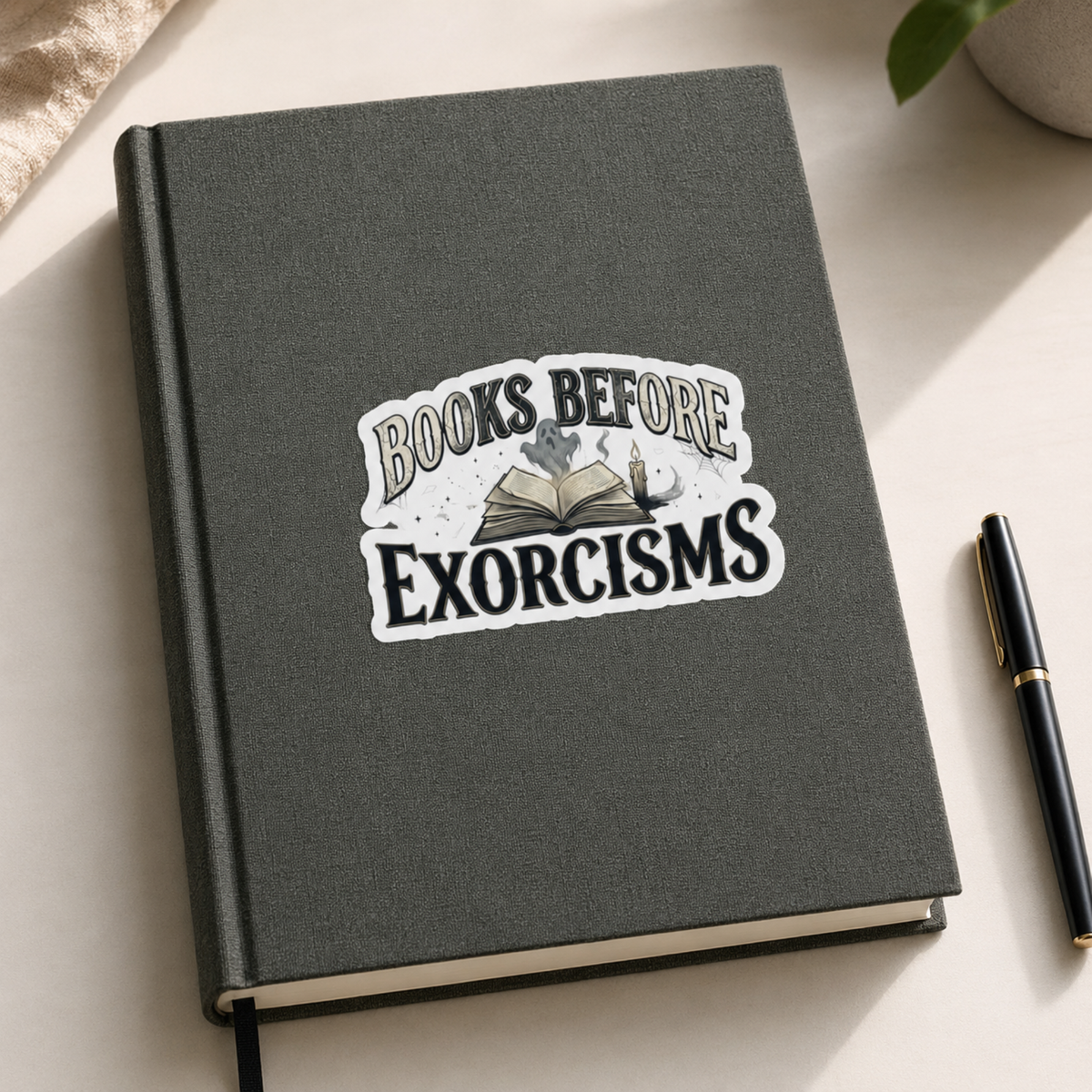 “Books Before Exorcisms” sticker with black lettering, an open book, gray ghost, candle, and cobweb, shown on a gray journal.